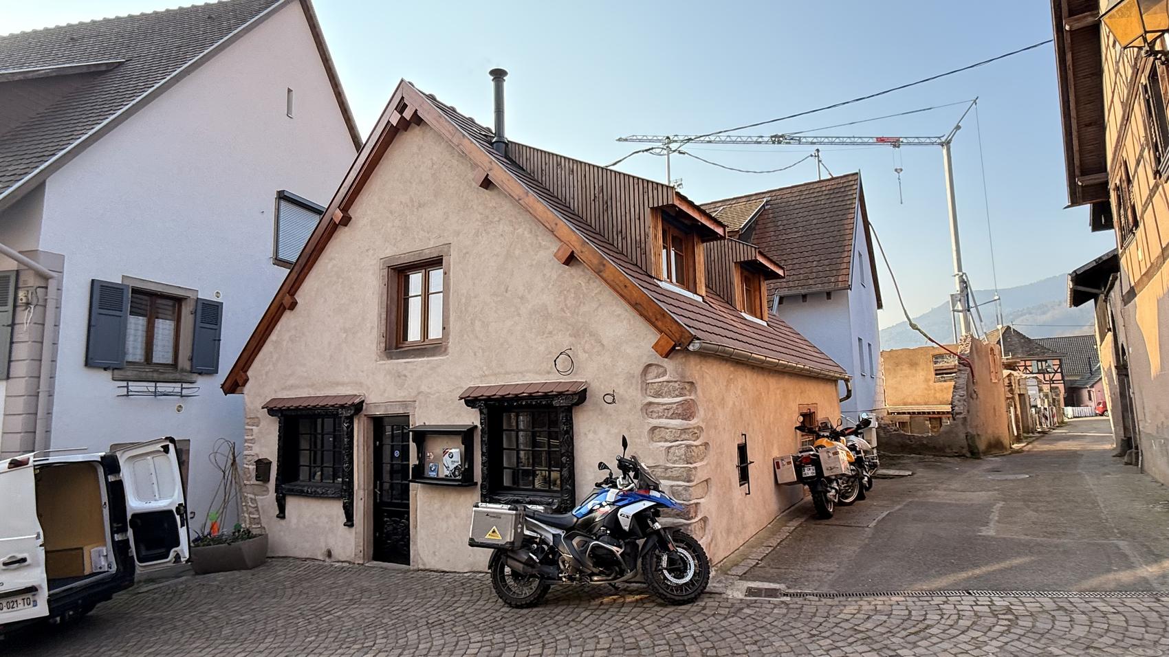 La façade du Schopf, une maison de pierre à l'angle d'une rue pavée d'Ottrott, deux motos garées devant